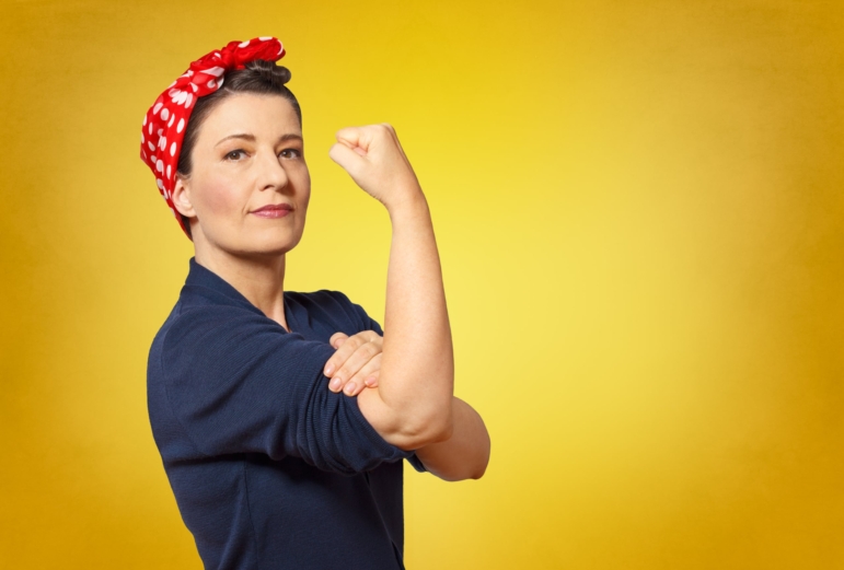 Self-confident middle aged woman wearing a red bandana and blue jumpsuit with rolled up sleeves with a clenched fist held up defiantly, a tribute to american icon Rosie the Riveter.